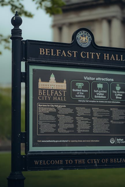 A black metal information sign for Belfast City Hall, featuring a large rectangular display with a rounded top and decorative finials on the posts. The sign includes the Belfast City Hall emblem at the top, a graphic of the historic building, and sections detailing visitor attractions such as guided tours, self-guided exhibitions, and the Bobbin Coffee Shop. The background shows a partly cloudy sky and some greenery, with the sign standing on a paved area. Catford Carpet Cleaning emphasizes thorough surface cleaning and sanitisation processes, ensuring maintained hygiene for public spaces like Belfast City Hall, consistent with domestic and commercial cleaning standards.
