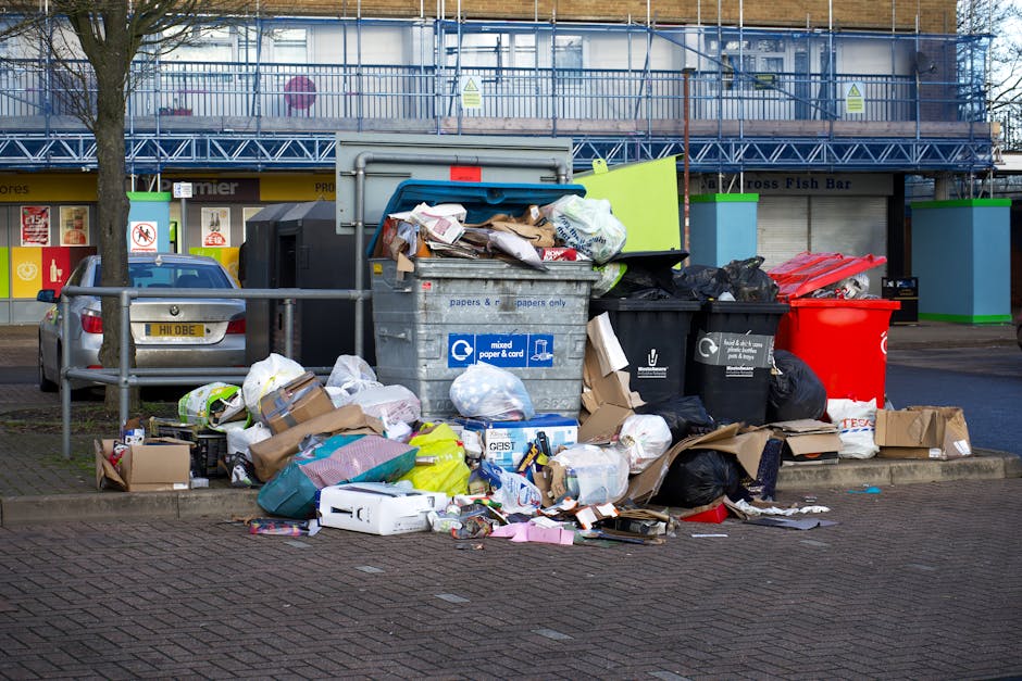 An outdoor waste collection area in Lewisham featuring large, overflowing rubbish bins and open recycling containers filled with mixed paper, cardboard, black plastic bags, and discarded packaging. The scene includes a paved sidewalk, a parked silver car on the left, and a metal railing separating the waste area from the pedestrian path. In the background, there is a building with a blue metal fire escape, storefronts, and a yellow sign for a fish bar. The waste appears cluttered, with items spilling onto the ground and surrounding the containers, indicating an unkempt state. Natural daylight illuminates the scene, highlighting the varied materials and the disorderly arrangement of the garbage, which suggests the need for proper waste management and cleaning services in accordance with local municipal regulations.