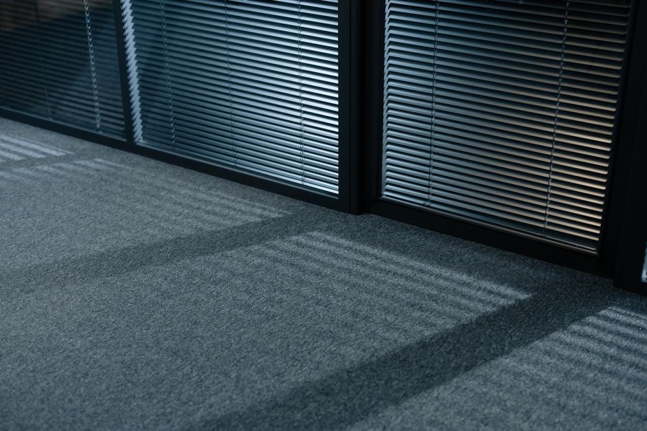 Close-up view of a modern office interior featuring a dark metal louvered radiator cover casting shadows on a clean, light gray carpeted floor. The carpet has a uniform, low-pile texture with no visible stains or debris, indicating recent professional deep cleaning. The room is illuminated by natural light filtering through horizontal window blinds, creating a pattern of light and shadow on the floor. The scene reflects a well-maintained workspace emphasizing surface cleaning and hygiene, consistent with the office carpet cleaning services offered by Catford Carpet Cleaning. The overall appearance suggests meticulous sanitisation and ongoing maintenance to ensure a hygienic environment.