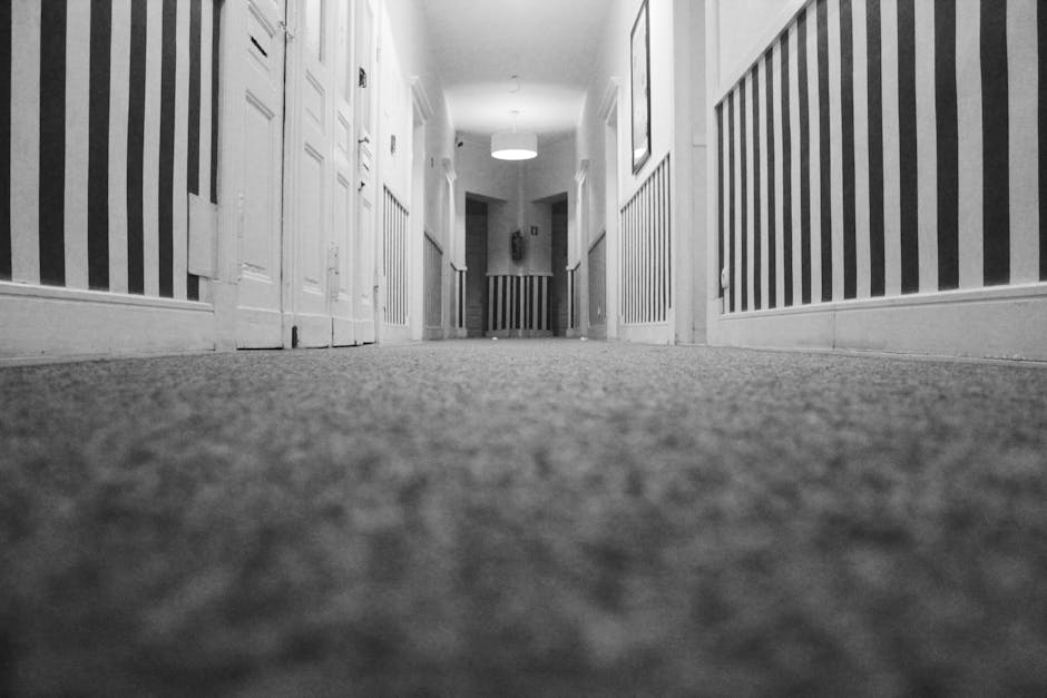 A long, narrow corridor in a commercial building with a clean, well-maintained appearance. The ceiling is fitted with evenly spaced, recessed lighting fixtures that provide uniform illumination. The walls are painted white and feature several doorways with dark frames, likely leading to individual offices or rooms. On the left side, there is a rectangular ventilation grille visible near the ceiling. The corridor is covered with a textured, patterned carpet in shades of grey, appearing fresh and free of dirt or stains. The overall scene reflects a high-standard of cleanliness maintained by [COMPANY_NAME], emphasizing surface cleaning and hygiene suitable for office environments. The space is unoccupied, showcasing a professional, sanitised setting ready for occupants or visitors.