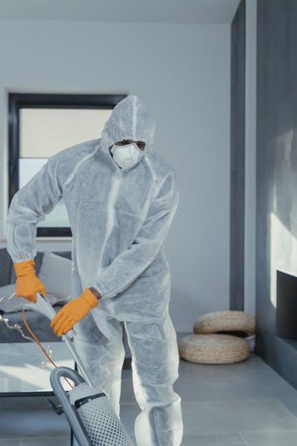 A professional cleaner dressed in a white protective suit, mask, and orange gloves is performing surface cleaning with a spray bottle and vacuum cleaner in a modern, well-lit living room. The room features large windows allowing natural light, sleek gray tile flooring, and minimal decor including a pair of round, beige cushions placed on the floor beside a gray wall. The cleaning process appears thorough, contributing to hygiene and maintaining the cleanliness of the space, exemplifying domestic cleaning services offered by Catford Carpet Cleaning in Rushey Green SE6. The setting emphasizes a high standard of deep cleaning and sanitisation, suitable for both residential and commercial environments.