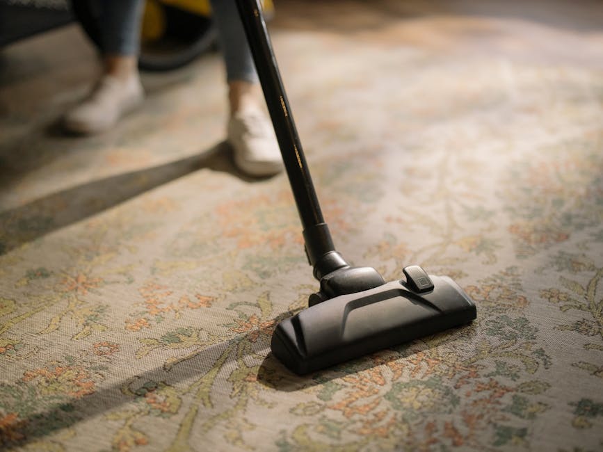 A professional cleaner dressed in a white protective suit, mask, and orange gloves is performing surface cleaning with a spray bottle and vacuum cleaner in a modern, well-lit living room. The room features large windows allowing natural light, sleek gray tile flooring, and minimal decor including a pair of round, beige cushions placed on the floor beside a gray wall. The cleaning process appears thorough, contributing to hygiene and maintaining the cleanliness of the space, exemplifying domestic cleaning services offered by Catford Carpet Cleaning in Rushey Green SE6. The setting emphasizes a high standard of deep cleaning and sanitisation, suitable for both residential and commercial environments.