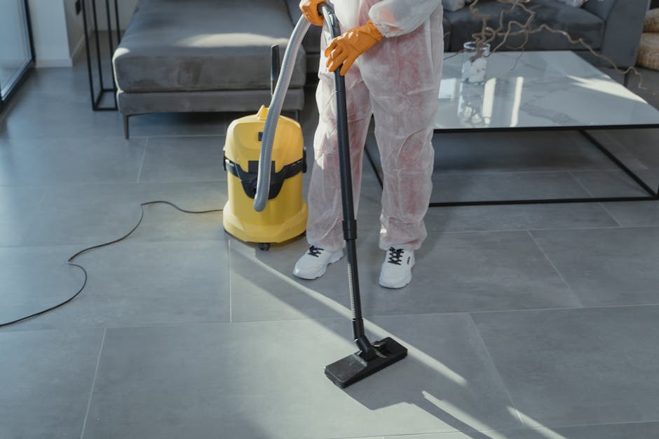 A person wearing white protective clothing, gloves, and sneakers is using a vacuum and a floor cleaning attachment to deep clean large, square, light grey ceramic tiles in a modern living room. The vacuum cleaner, which is yellow with a black handle, is positioned nearby on the polished tile floor. The room features a glass coffee table with a metal frame, grey upholstered sofas, and natural light streaming in through a nearby window, highlighting the spotless, dust-free surfaces. The scene depicts professional surface cleaning and sanitisation services provided by Catford Carpet Cleaning for domestic spaces, ensuring hygiene and pristine conditions in the living area.